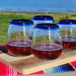 May include: Four stemless wine glasses with a blue rim, filled with red liquid, sit on a wooden cutting board. The glasses are on a red, white, and blue striped blanket, with a blurred outdoor background.