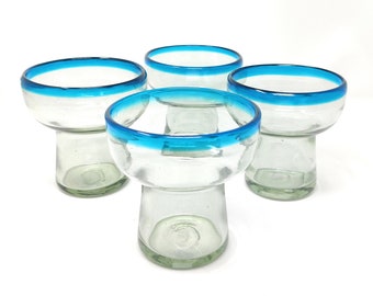 May include: Four clear glass drinking glasses with a blue rim. The glasses are shaped like a traditional Mexican shot glass with a wide bowl and a narrow base.