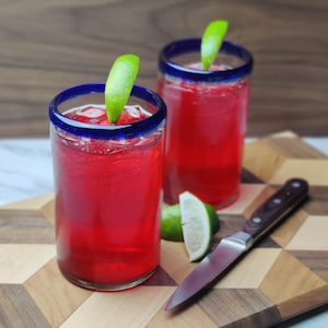 May include: Two tall glasses with blue rims filled with a bright red beverage and garnished with lime wedges. The glasses are on a wooden cutting board with a silver knife.