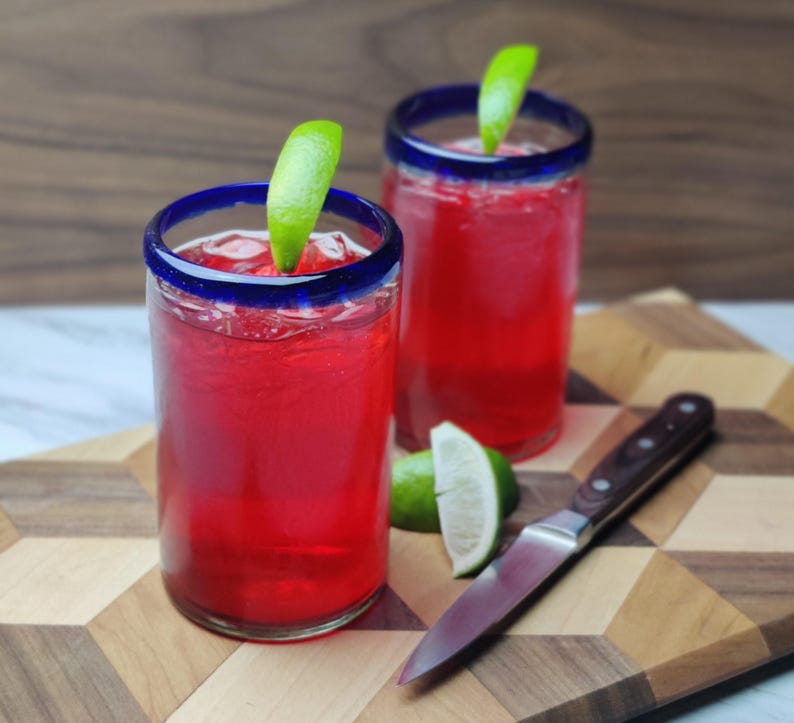 May include: Two tall glasses with blue rims filled with a bright red beverage and garnished with lime wedges. The glasses are on a wooden cutting board with a silver knife.