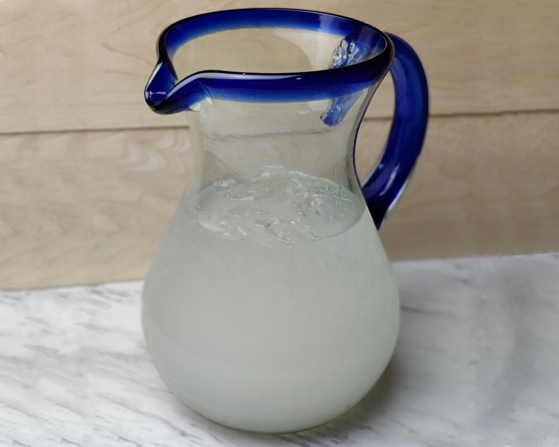 May include: A clear glass pitcher with a blue rim and handle, filled with a white liquid and ice.