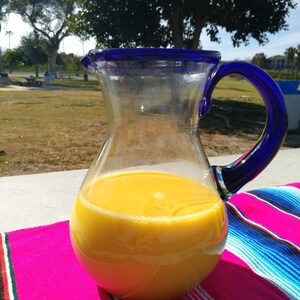 May include: A glass pitcher with a blue rim and handle filled with a yellow liquid. The pitcher is sitting on a colorful striped blanket.
