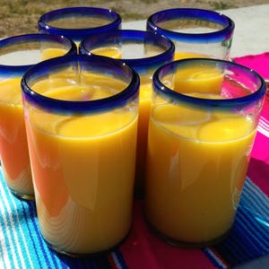 May include: Six glasses of orange juice in blue rimmed glasses on a colorful striped table cloth.