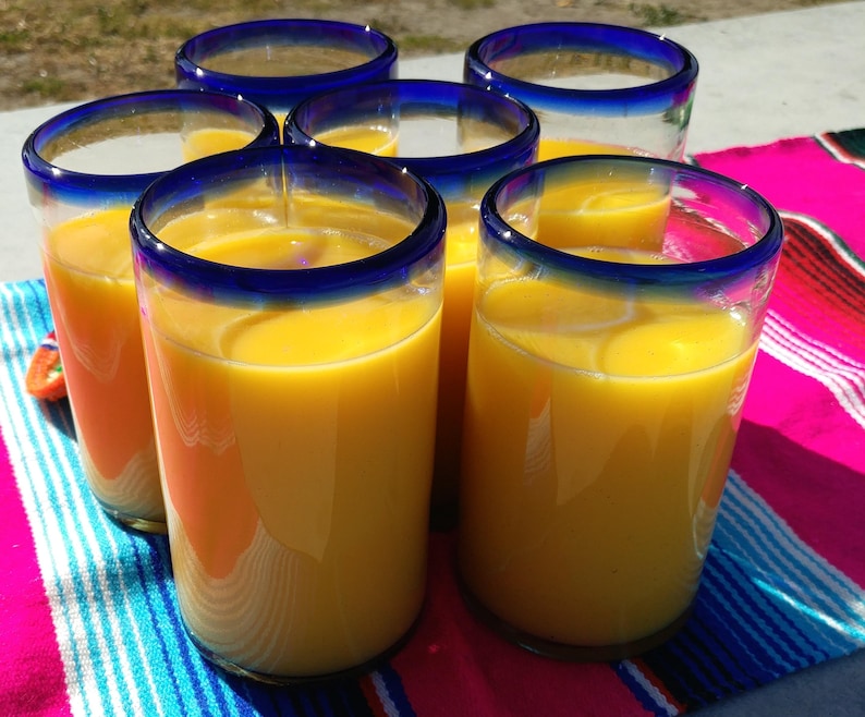 May include: Six glasses of orange juice in blue rimmed glasses on a colorful striped table cloth.