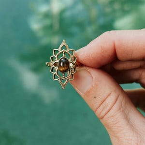 K&ouml;nnte beinhalten: Ein goldfarbener Ring mit einem komplizierten, symmetrischen, durchbrochenen Metallrahmen in Form einer Lotusbl&uuml;te. In der Mitte der dekorativen Metallarbeit befindet sich ein kleiner, glatter, ovaler brauner Edelstein mit einem dezenten Glanz.