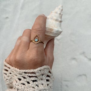 May include: A gold-colored ring with a teardrop-shaped design, featuring a small, round, light-colored stone. The ring is worn on a finger, and the hand is holding a white seashell. The sleeve of a cream-colored crocheted sweater is visible.