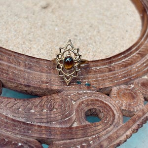 K&ouml;nnte beinhalten: Ein goldfarbener Ring mit einem ovalen, braunen Tigerauge-Edelstein, der in der Mitte eines dekorativen, symmetrischen Metallrahmens sitzt, mit spitzen, bl&uuml;tenblattartigen Aussparungen, die sich vertikal &uuml;ber und unter dem Stein erstrecken.