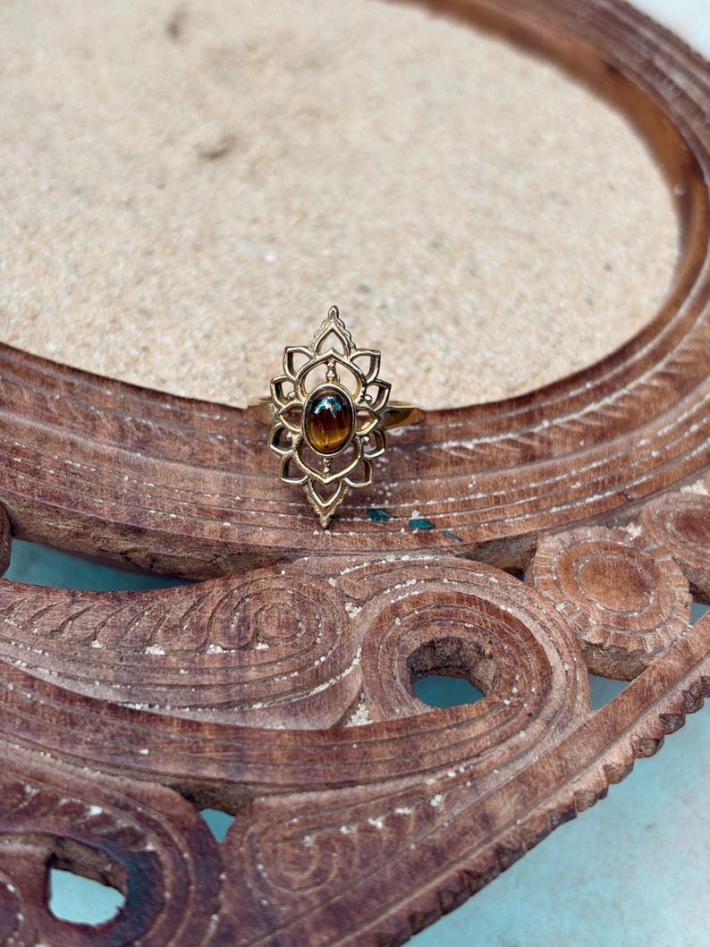 K&ouml;nnte beinhalten: Ein goldfarbener Ring mit einem ovalen, braunen Tigerauge-Edelstein, der in der Mitte eines dekorativen, symmetrischen Metallrahmens sitzt, mit spitzen, bl&uuml;tenblattartigen Aussparungen, die sich vertikal &uuml;ber und unter dem Stein erstrecken.