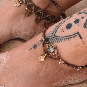 May include: Two brown ankle bracelets with gold accents. One bracelet features a small white seashell charm and the other has a larger charm with a moonstone.