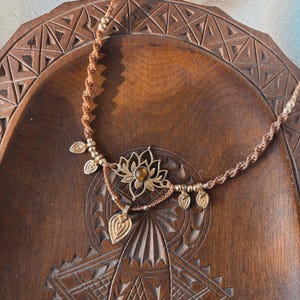 May include: A brown macrame cord necklace featuring a central gold-toned lotus flower pendant with a small amber-colored stone at its center, accented by five small gold-toned leaf-shaped charms and metallic beads along the braided cord.