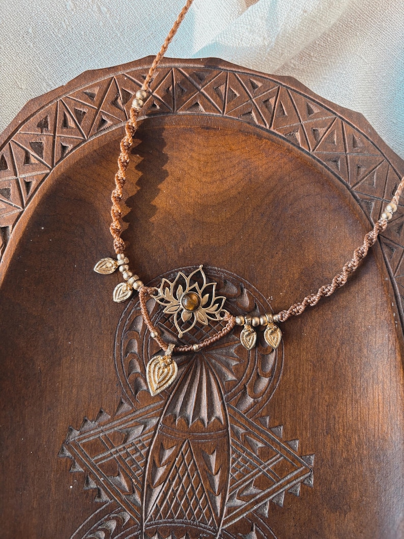 May include: A brown macrame cord necklace featuring a central gold-toned lotus flower pendant with a small amber-colored stone at its center, accented by five small gold-toned leaf-shaped charms and metallic beads along the braided cord.