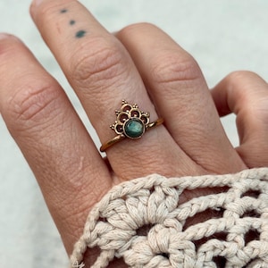 May include: A gold-colored ring with a crown-like design and a small, round, green gemstone. The ring is worn on a finger, and the hand is adorned with a crocheted accessory.