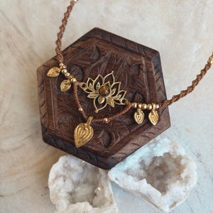 May include: A brown cord necklace featuring a central gold-toned lotus flower pendant with a small brown gemstone at its center. The necklace is adorned with several small gold-toned beads and dangling leaf-shaped charms along the cord.