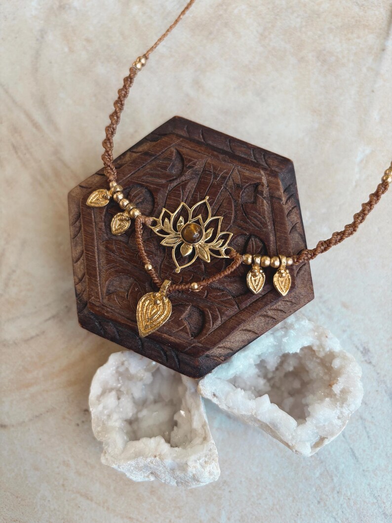 May include: A brown cord necklace featuring a central gold-toned lotus flower pendant with a small brown gemstone at its center. The necklace is adorned with several small gold-toned beads and dangling leaf-shaped charms along the cord.