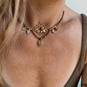 Puede incluir: Un collar tipo gargantilla de macramé marrón con un colgante central de flor de loto dorada con una piedra marrón oscuro. El collar está decorado con pequeñas cuentas doradas, dijes en forma de hoja y un colgante dorado en forma de lágrima.