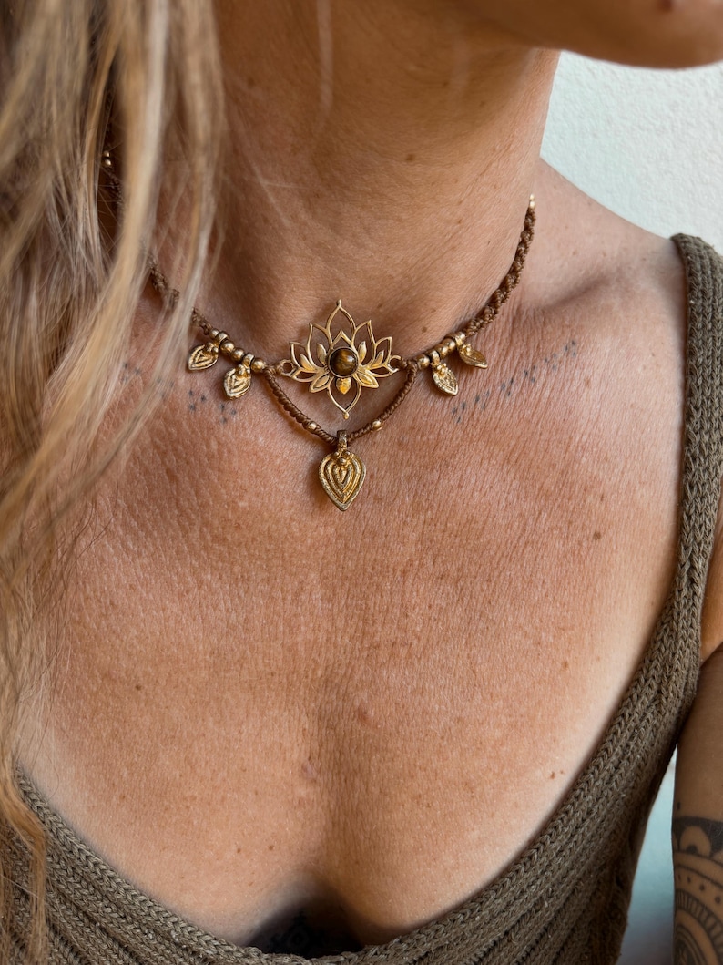 May include: A brown macrame choker necklace featuring a central gold-toned lotus flower pendant with a dark brown stone inlay. The necklace is accented with small gold-toned beads and leaf-shaped charms, with a teardrop-shaped gold-toned pendant hanging below.