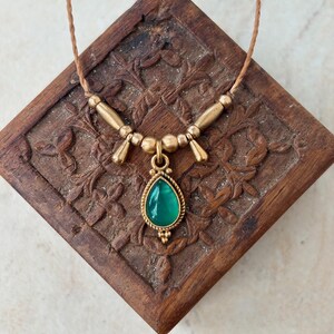 May include: A necklace featuring a teardrop-shaped green stone pendant set in a gold-toned metal frame. The pendant hangs from a thin brown cord accented with gold-toned cylindrical and spherical beads arranged symmetrically on each side.