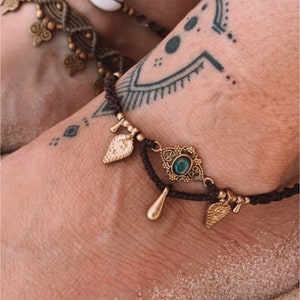 May include: A brown ankle bracelet with gold charms, including a teardrop, a leaf, and a decorative charm with a green gemstone. The bracelet is worn on a person's ankle with a tattoo.