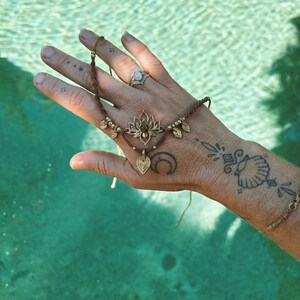 May include: A brown macrame bracelet featuring a central gold-toned lotus flower charm with a small embedded stone, accented by metallic beads and a dangling leaf-shaped pendant, secured with thin adjustable cords.