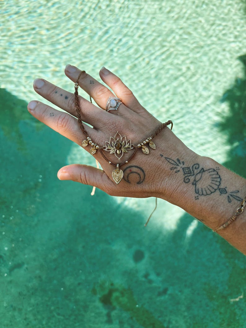 May include: A brown macrame bracelet featuring a central gold-toned lotus flower charm with a small embedded stone, accented by metallic beads and a dangling leaf-shaped pendant, secured with thin adjustable cords.