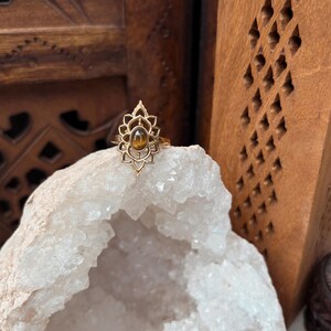 K&ouml;nnte beinhalten: Ein goldfarbener Ring mit einem komplizierten, symmetrischen, von Lotusbl&uuml;ten inspirierten Metalldesign, das einen zentralen, ovalen, braunen Tigeraugen-Edelstein umgibt. Der Ring liegt auf einer gro&szlig;en, gezackten, wei&szlig;en Kristallgeode.
