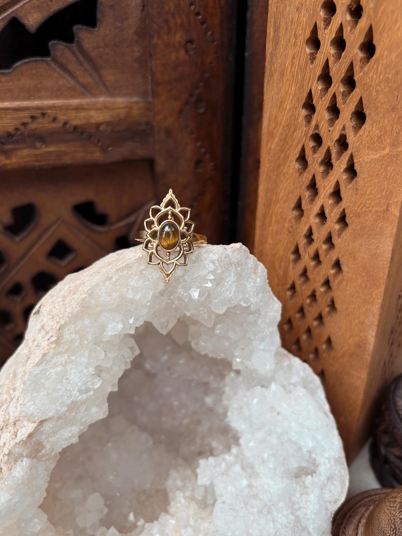 K&ouml;nnte beinhalten: Ein goldfarbener Ring mit einem komplizierten, symmetrischen, von Lotusbl&uuml;ten inspirierten Metalldesign, das einen zentralen, ovalen, braunen Tigeraugen-Edelstein umgibt. Der Ring liegt auf einer gro&szlig;en, gezackten, wei&szlig;en Kristallgeode.
