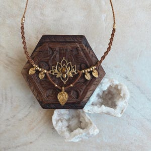 May include: A brown macrame necklace featuring a central gold-toned lotus flower pendant with a brown gemstone at its center. The necklace includes small gold-toned beads and several small gold-toned leaf-shaped charms hanging from the cord.
