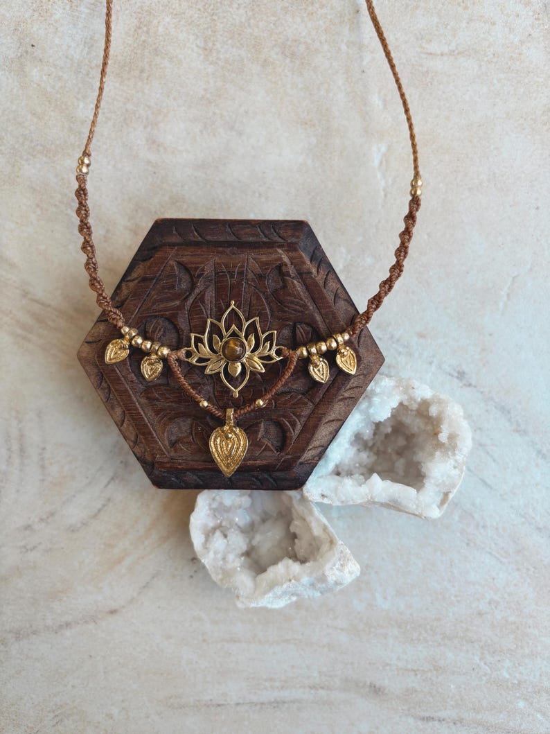 May include: A brown macrame necklace featuring a central gold-toned lotus flower pendant with a brown gemstone at its center. The necklace includes small gold-toned beads and several small gold-toned leaf-shaped charms hanging from the cord.