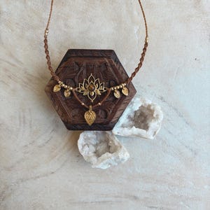 May include: A brown cord necklace featuring a central gold-toned lotus flower pendant with a small brown stone, accented by four small gold-toned leaf charms and a single dangling heart-shaped charm, all suspended on a braided cord.