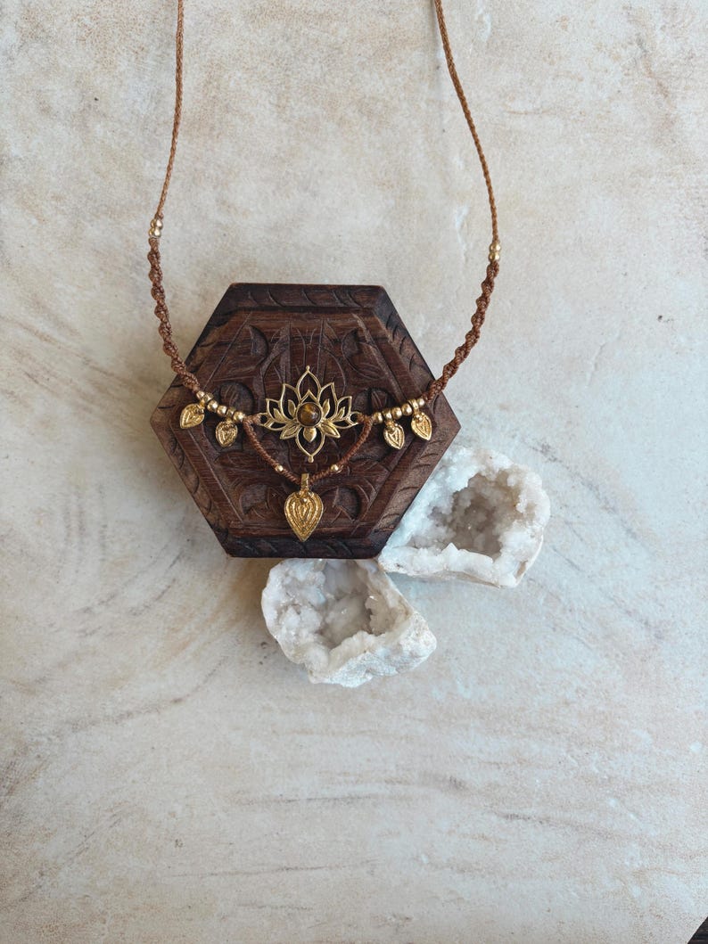 May include: A brown cord necklace featuring a central gold-toned lotus flower pendant with a small brown stone, accented by four small gold-toned leaf charms and a single dangling heart-shaped charm, all suspended on a braided cord.