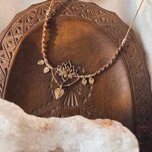 May include: A brown macrame cord necklace featuring a central gold-toned lotus flower pendant with a small amber-colored stone at its center. The necklace is adorned with several small, dangling gold-toned leaf-shaped charms along the cord.