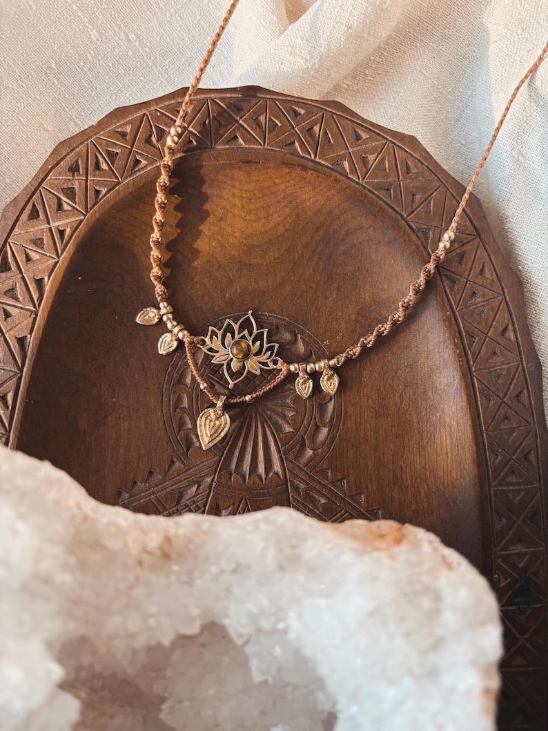 May include: A brown macrame cord necklace featuring a central gold-toned lotus flower pendant with a small amber-colored stone at its center. The necklace is adorned with several small, dangling gold-toned leaf-shaped charms along the cord.