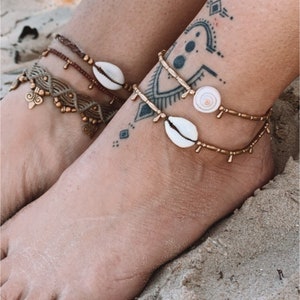 May include: Close-up of two ankles with anklets on a sandy beach. One ankle has a brown macrame anklet with gold accents and a seashell. The other has a gold beaded anklet with a seashell.