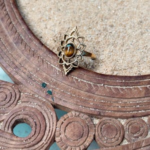 K&ouml;nnte beinhalten: Ein goldfarbener Ring mit einem l&auml;nglichen, durchbrochenen Metallrahmen und einem zentralen, ovalen braunen Tigerauge-Edelstein. Die Fassung weist ein symmetrisches, bl&uuml;tenblattartiges Design um den Stein herum auf.