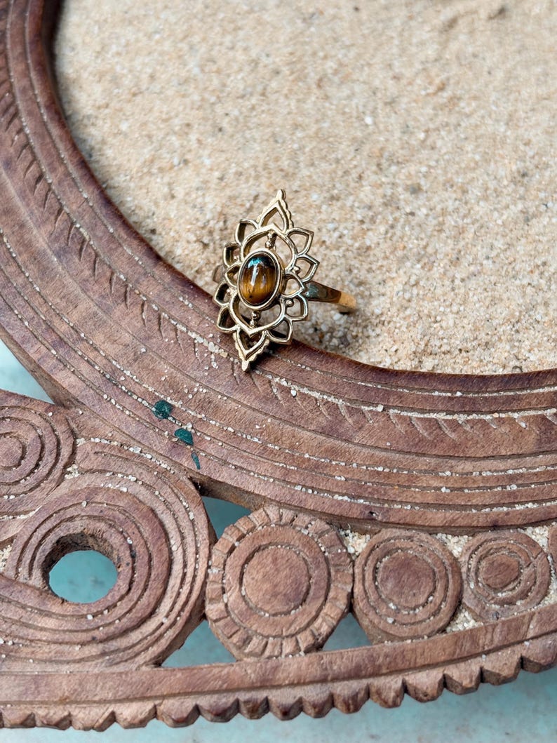 K&ouml;nnte beinhalten: Ein goldfarbener Ring mit einem l&auml;nglichen, durchbrochenen Metallrahmen und einem zentralen, ovalen braunen Tigerauge-Edelstein. Die Fassung weist ein symmetrisches, bl&uuml;tenblattartiges Design um den Stein herum auf.