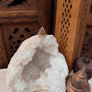 K&ouml;nnte beinhalten: Ein goldfarbener Ring mit einem komplizierten, spitz zulaufenden, bl&uuml;tenblattartigen Metallrahmen, der einen kleinen, ovalen, braunen Tigeraugen-Edelstein umschlie&szlig;t. Der Ring liegt auf einer gro&szlig;en, hohlen, wei&szlig;en Kristallgeode.