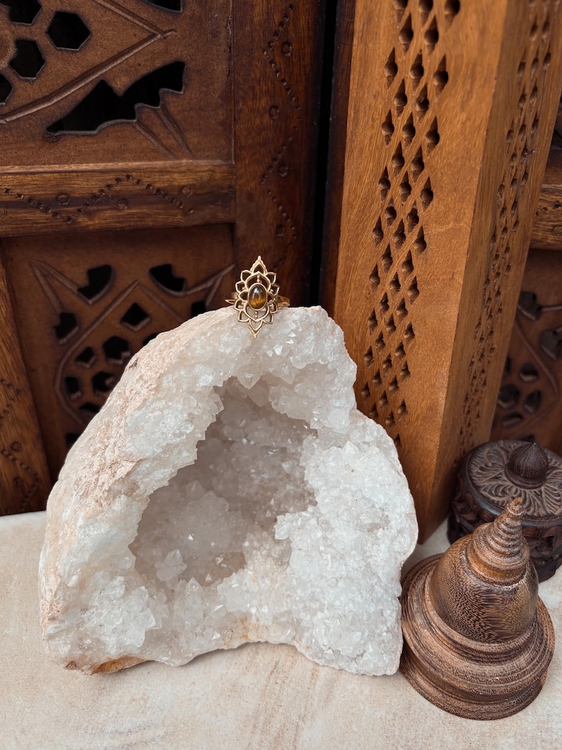 K&ouml;nnte beinhalten: Ein goldfarbener Ring mit einem komplizierten, spitz zulaufenden, bl&uuml;tenblattartigen Metallrahmen, der einen kleinen, ovalen, braunen Tigeraugen-Edelstein umschlie&szlig;t. Der Ring liegt auf einer gro&szlig;en, hohlen, wei&szlig;en Kristallgeode.