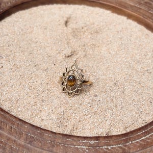 K&ouml;nnte beinhalten: Ein goldfarbener Ring mit einem ovalen, braunen Tigerauge-Edelstein, der in der Mitte eines dekorativen, symmetrischen Metallrahmens sitzt, welcher mit spitz zulaufenden, bl&uuml;tenblattartigen Aussparungen um den Stein herum versehen ist.