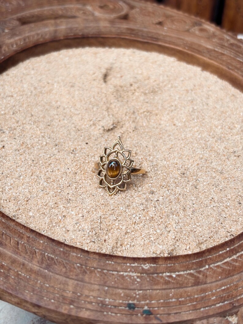 K&ouml;nnte beinhalten: Ein goldfarbener Ring mit einem ovalen, braunen Tigerauge-Edelstein, der in der Mitte eines dekorativen, symmetrischen Metallrahmens sitzt, welcher mit spitz zulaufenden, bl&uuml;tenblattartigen Aussparungen um den Stein herum versehen ist.