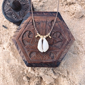 May include: A brown cord necklace with a white seashell pendant and gold beads. The necklace is displayed on a wooden box with intricate carvings.
