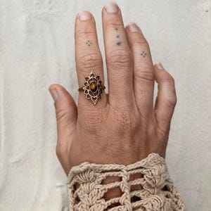 K&ouml;nnte beinhalten: Ein goldfarbener Ring mit einem ovalen, braunen Tigerauge-Edelstein, der in einen dekorativen, spitz zulaufenden Metallrahmen gefasst ist. Der Ring wird am Mittelfinger getragen und zeigt ein detailliertes, symmetrisches Design mit kleinen Aussparungen.