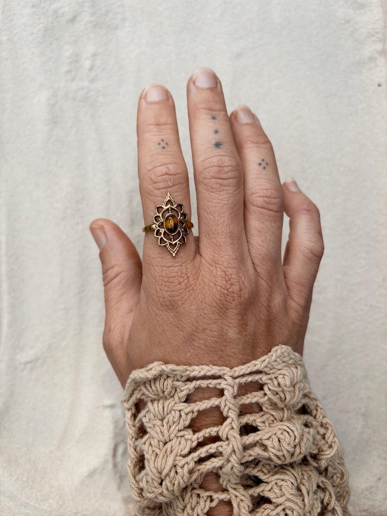K&ouml;nnte beinhalten: Ein goldfarbener Ring mit einem ovalen, braunen Tigerauge-Edelstein, der in einen dekorativen, spitz zulaufenden Metallrahmen gefasst ist. Der Ring wird am Mittelfinger getragen und zeigt ein detailliertes, symmetrisches Design mit kleinen Aussparungen.
