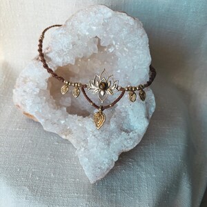 May include: A brown macrame cord necklace featuring a central gold-toned lotus flower pendant with a small brown gemstone at its center. The necklace is adorned with several small gold-toned metal leaf-shaped charms hanging from the cord.