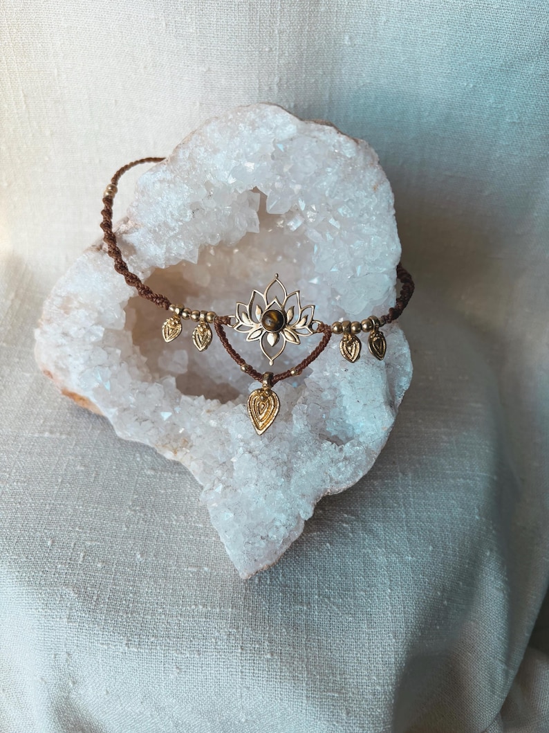 May include: A brown macrame cord necklace featuring a central gold-toned lotus flower pendant with a small brown gemstone at its center. The necklace is adorned with several small gold-toned metal leaf-shaped charms hanging from the cord.