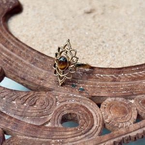 K&ouml;nnte beinhalten: Ein goldfarbener Ring mit einem ovalen, braunen Tigerauge-Edelstein, der in der Mitte eines komplizierten, durchbrochenen Metallrahmens sitzt, welcher an eine stilisierte Lotusblume mit spitz zulaufenden Bl&uuml;tenbl&auml;ttern erinnert.