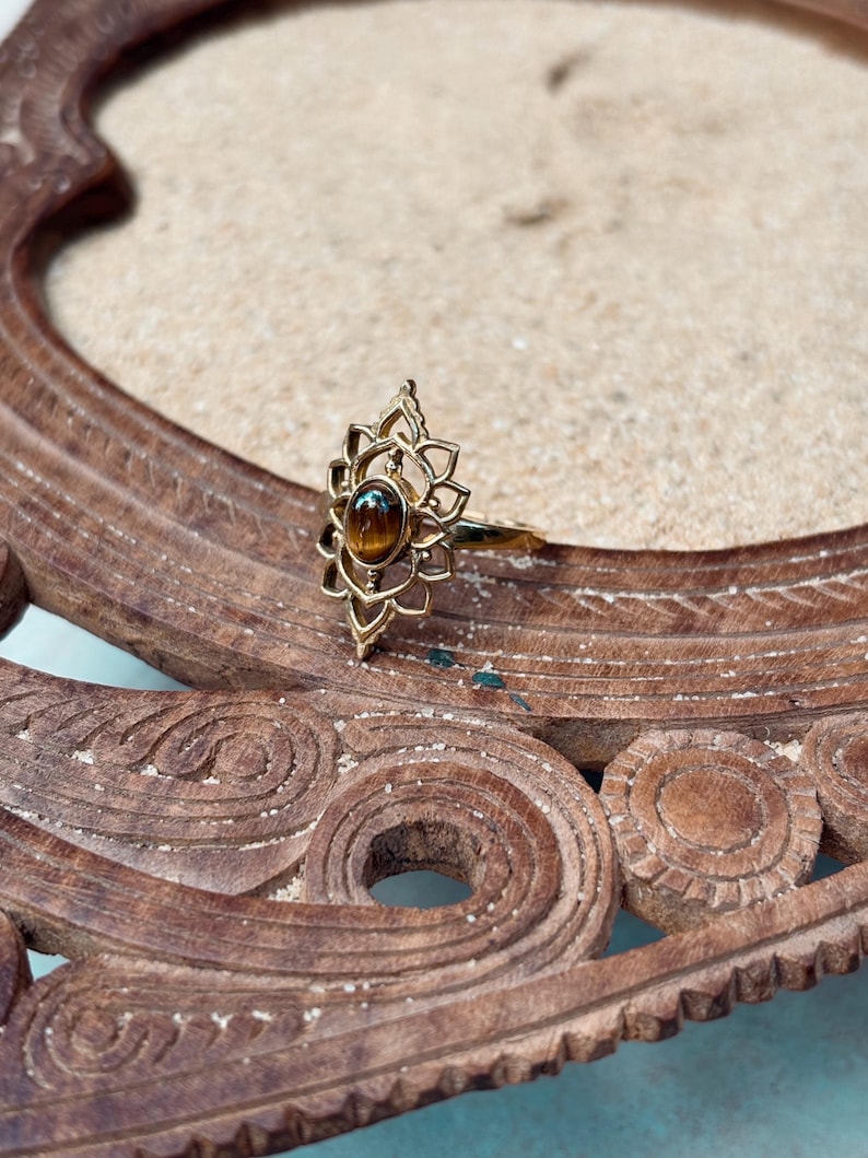K&ouml;nnte beinhalten: Ein goldfarbener Ring mit einem ovalen, braunen Tigerauge-Edelstein, der in der Mitte eines komplizierten, durchbrochenen Metallrahmens sitzt, welcher an eine stilisierte Lotusblume mit spitz zulaufenden Bl&uuml;tenbl&auml;ttern erinnert.