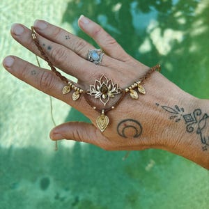 May include: A brown cord bracelet featuring a central gold-toned lotus flower charm with a small brown stone at its center. The bracelet includes gold-toned metal beads and three dangling leaf-shaped charms, with adjustable cord ends.