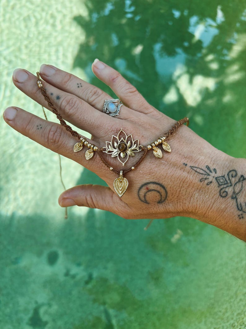 May include: A brown cord bracelet featuring a central gold-toned lotus flower charm with a small brown stone at its center. The bracelet includes gold-toned metal beads and three dangling leaf-shaped charms, with adjustable cord ends.