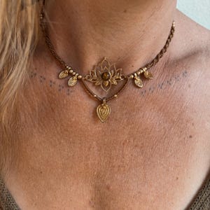 May include: A brown macrame choker necklace featuring a central gold-toned lotus flower charm with a round brown gemstone at its center. The necklace is accented with small gold-toned beads and four dangling leaf-shaped gold-toned metal charms.