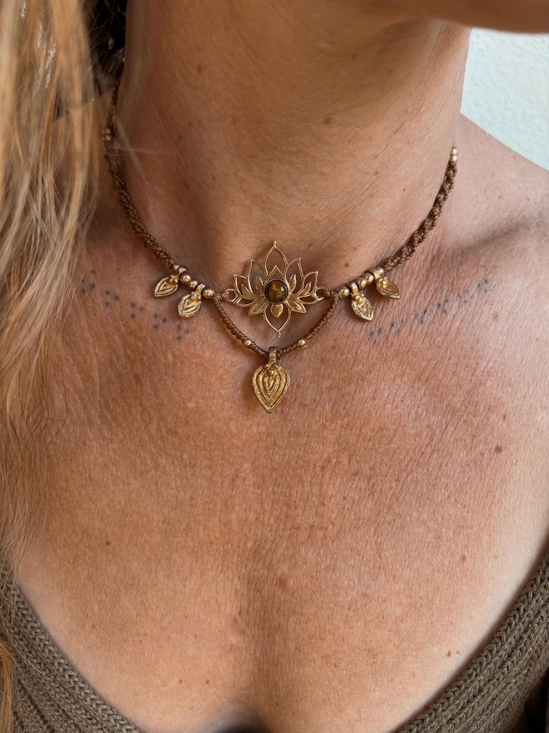 May include: A brown macrame choker necklace featuring a central gold-toned lotus flower charm with a round brown gemstone at its center. The necklace is accented with small gold-toned beads and four dangling leaf-shaped gold-toned metal charms.